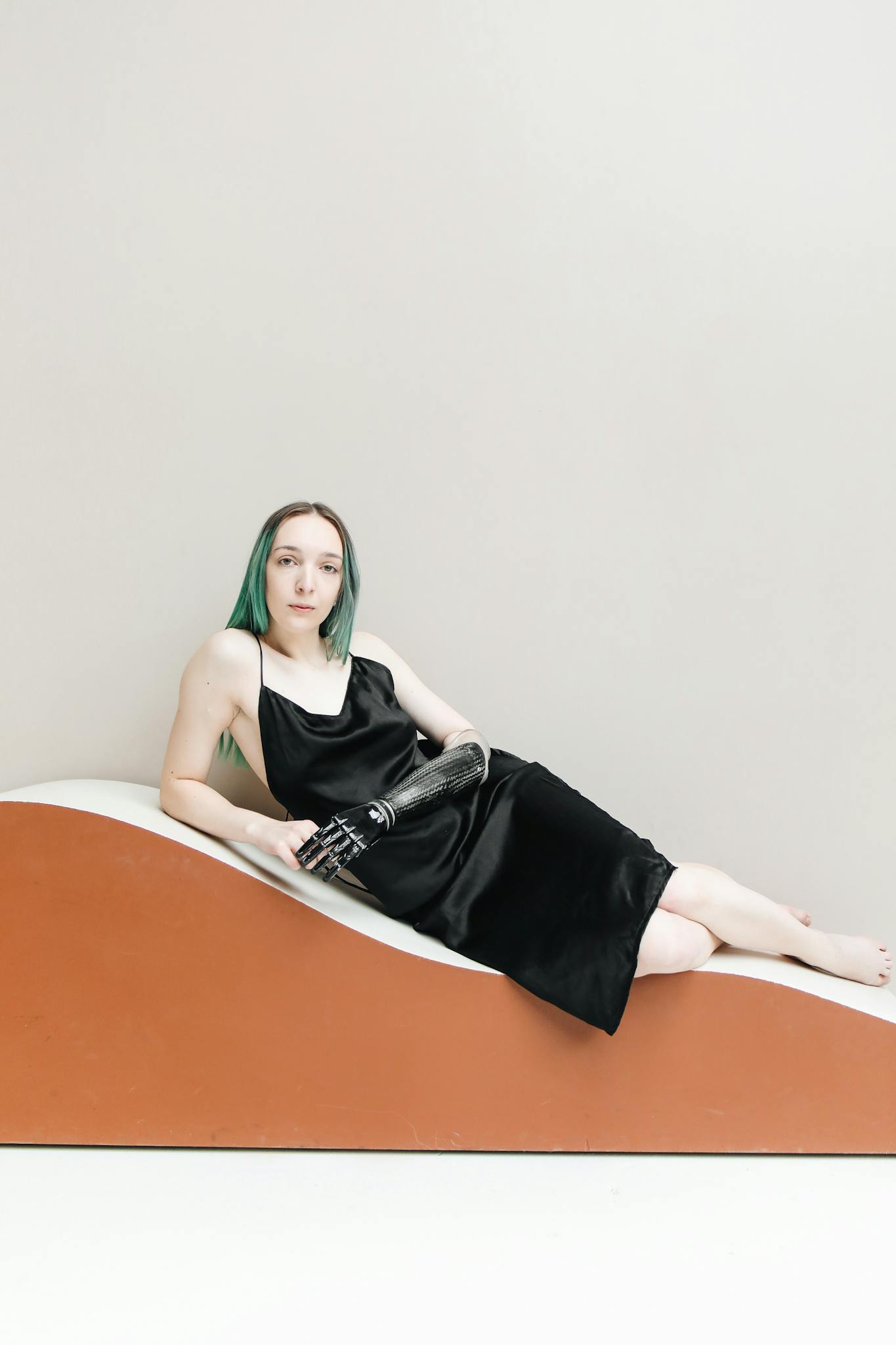 Caucasian woman with green hair reclines in a modern setting, showcasing a sleek prosthetic arm.
